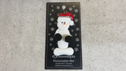 Ornament of white snowman in red hat holding black cheerleader pom-poms, shown on backing card and Christmas tree.