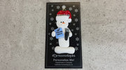 Ornament of white snowman in red hat holding a microphone and blue music book, shown on backing card and Christmas tree.