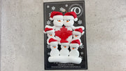 Family ornament with 7 white snowmen in red hats, gathered around a red maple leaf, shown on backing card and Christmas tree.