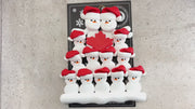 Family ornament with 13 white snowmen in red hats, gathered around a red maple leaf, shown on backing card and Christmas tree.