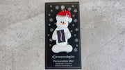 Ornament of white snowman in red hat holding black and white x-ray of a spine, shown on backing card and Christmas tree.