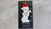 Ornament of white snowman in red hat holding a white diploma, shown on backing card and Christmas tree.