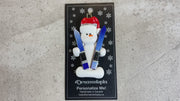Ornament of white snowman in red hat holding a pair of vibrant skis, shown on backing card and Christmas tree.