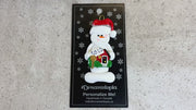 Ornament of white snowman in red hat holding a sign with a house saying “sold”, shown on backing card and Christmas tree.