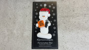 Ornament of white snowman in red hat holding a microphone and orange music book, shown on backing card and Christmas tree.