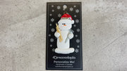 Ornament of white snowman in red hat holding a piece of wood and a screw, shown on backing card and Christmas tree.