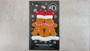 Family ornament with 2 brown gingerbread figures in red Christmas hats, shown on backing card and Christmas tree.