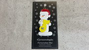 Ornament of white snowman in red hat holding a yellow music note, shown on backing card and Christmas tree.