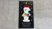 Ornament of white snowman in red hat holding various pills and a mortar and pestle, shown on backing card and Christmas tree.