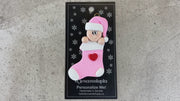 Ornament with infant head in a pink hat poking out of a pink stocking, shown on backing card and Christmas tree.
