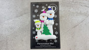 Family ornament with 3 white snowmen in bright scarves, with green Christmas tree, shown on backing card and Christmas tree.