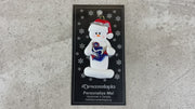 Ornament of white snowman in red hat holding a plastic anchor, shown on backing card and Christmas tree.