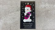 Ornament of white snowman in red hat holding a vibrant snowboard, shown on backing card and Christmas tree.