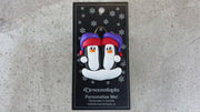 A couple’s ornament of two penguins in colourful hats, shown on backing card and Christmas tree.