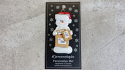 Ornament of white snowman in red hat holding several parcels, shown on backing card and Christmas tree.