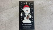 Ornament of white snowman in red hat holding a hunting rifle and pair of binoculars, shown on backing card and Christmas tree.
