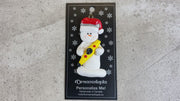 Ornament of white snowman in red hat holding a yellow kayak, shown on backing card and Christmas tree.