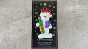 Ornament of white snowman in red hat holding a cluster of grapes and a wine bottle, shown on backing card and Christmas tree.