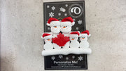 Family ornament with 5 white snowmen in red hats, gathered around a red maple leaf, shown on backing card and Christmas tree.