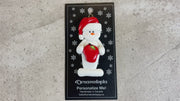 Ornament of white snowman in red hat holding a shiny red apple, shown on backing card and Christmas tree.