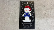 Ornament of white snowman in red hat holding a blue video game controller, shown on backing card and Christmas tree.