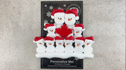 Family ornament with 10 white snowmen in red hats, gathered around a red maple leaf, shown on backing card and Christmas tree.
