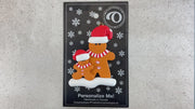 Family ornament with 1 large and 1 small brown gingerbread figure in red Christmas hats, shown on backing card and Christmas tree.