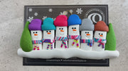 Family ornament with 6 snowmen in colourful scarves, hemmed by Christmas trees, shown on backing card and Christmas tree.