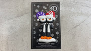 Family ornament with 2 black and white penguins in colourful festive hats, shown on backing card and Christmas tree.