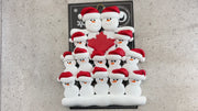 Family ornament with 14 white snowmen in red hats, gathered around a maple leaf, shown on backing card and Christmas tree.