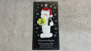 Ornament of white snowman in red hat holding a microphone and green music book, shown on backing card and Christmas tree.