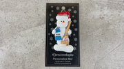 Ornament of white snowman in red hat holding a broom and bottle of cleaning solution, shown on backing card and Christmas tree.