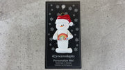 White snowman with small rainbow and red festive hat ornament, shown on backing card and Christmas tree.