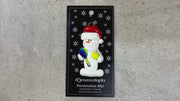 Ornament of white snowman in red hat holding a tennis paddle and ball, shown on backing card and Christmas tree.