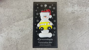 Ornament of white snowman in red hat holding a yellow school bus, shown on backing card and Christmas tree.