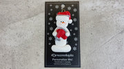 Ornament of white snowman in red hat holding a red airplane, shown on backing card and Christmas tree.