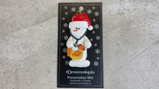 Ornament of white snowman in red hat holding chicken and stethoscope, shown on backing card and Christmas tree.