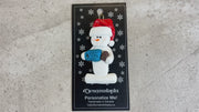 Ornament of white snowman in red hat holding colourful mug and coffee bean, shown on backing card and Christmas tree.
