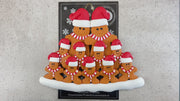 Family ornament with 11 brown gingerbread figures in red Christmas hats, shown on backing card and Christmas tree.