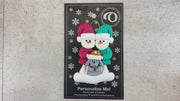 Ornament of snowman couple in red and green shirts and hats with grey dog in Christmas hat, shown on backing card and Christmas tree.