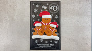 Family ornament with 1 large and 2 small brown gingerbread figures in red Christmas hats, shown on backing card and Christmas tree.