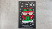 A couple’s ornament with two small red and green elves with a matching present, shown with backing card and Christmas tree.