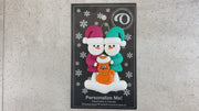 Ornament of snowman couple in red and green shirts and hats with a orange cat in Christmas hat, shown on backing card and Christmas tree.