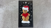Tan bear in red festive hat holding red heart ornament, shown on backing card and Christmas tree.