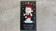 Ornament of white snowman in red hat holding a red tractor, shown on backing card and Christmas tree.