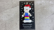 Ornament of white snowman in red hat holding an in-progress knitting project on knitting needles, shown on backing card and Christmas tree.