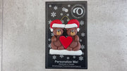 A couple’s ornament of two brown bears holding a red heart together, shown on backing card.