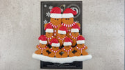 Family ornament with 9 brown gingerbread figures in red Christmas hats, shown on backing card and Christmas tree.