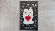 A couple’s ornament of two white snowmen holding a red heart together, shown on backing card and Christmas tree.