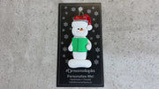 Ornament of white snowman in red hat holding a green book, shown on backing card and Christmas tree.